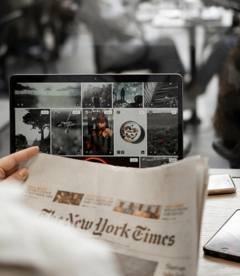 A person reading a newspaper while browsing images on a laptop, symbolizing remote work lifestyle.